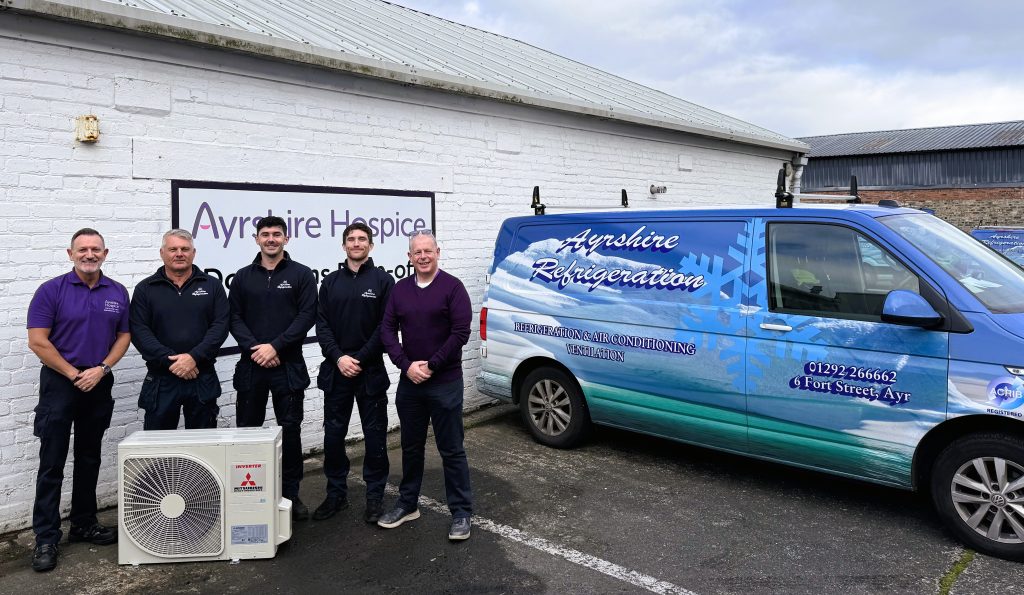 Five men standing outdoors in front of a white brick building. To the right of the men is a white outdoor air conditioning unit. Next to the men and the unit is a blue and white van with "Ayrshire Refrigeration" and "Refrigeration & Air Conditioning Ventilation" printed on its side.