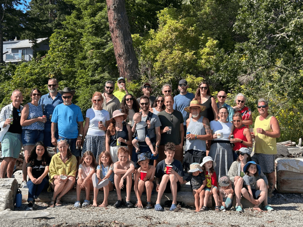 A large group of adults and children pose for a photo outdoors on a sunny day, with trees and a building in the background.