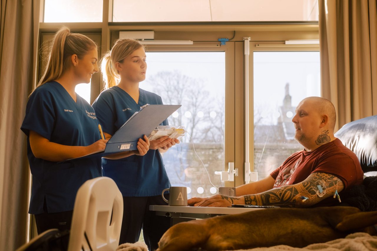 Two nurses in blue scrubs consult with a patient sitting in a blue chair with his hands on a table in front of him.