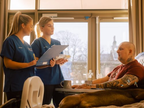 Two nurses in blue scrubs consult with a patient sitting in a blue chair with his hands on a table in front of him.