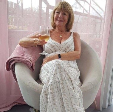 Margaret Cassidy smiling in a white patterned dress relaxing in a modern chair holding a glass of wine.