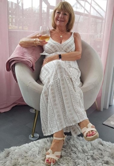 Margaret Cassidy smiling in a white patterned dress relaxing in a modern chair holding a glass of wine.