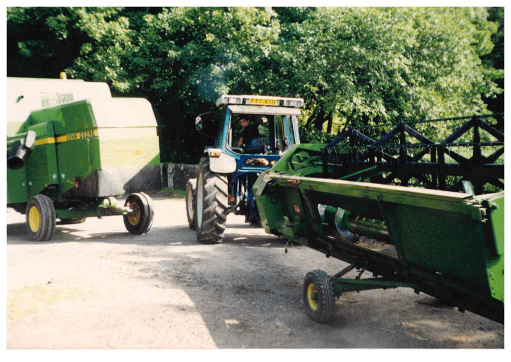 Alan driving tractor towing green farm machinery on a gravel road beside trees.