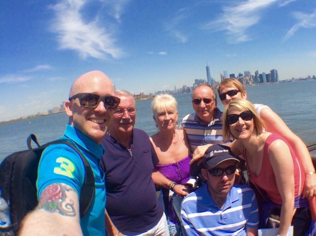 A group of seven adults posing for a selfie outdoors on a sunny day with the city skyline and water in the background.
